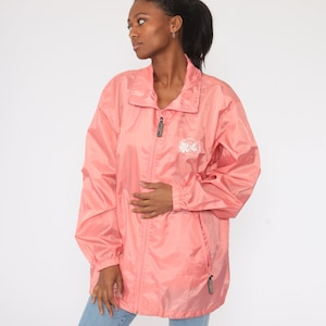 May include: A coral-colored, zip-up windbreaker jacket with a stand-up collar. The jacket has a white logo on the chest and a zipper pull. The jacket is made of a lightweight, shiny material.