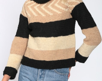 May include: A knitted sweater with a chevron pattern on the upper chest and horizontal stripes in black, tan, and cream. The sweater has long sleeves and a wide neckline. The model is wearing blue jeans.