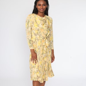 May include: A vintage yellow floral dress with long sleeves and a pleated skirt. The dress features a cowl neckline and a tie at the waist. The floral pattern includes yellow, white, and green flowers. The dress is knee-length.