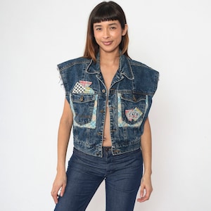 Op de afbeelding: Een blauwe denimvest met patchwork details en een geruit patroon. De vest heeft een knoopsluiting en twee zakken aan de voorkant. De vest is mouwloos en heeft een verweerde look.