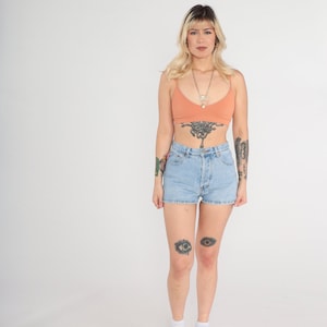 Op de afbeelding: Een vrouw draagt een lichtoranje crop top met een zilveren ketting en een hoge taille lichtblauwe denim short. Ze heeft tatoeages op haar armen, borst en knieën.