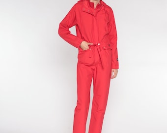 May include: A red hooded jumpsuit with a drawstring waist and long sleeves. The jumpsuit has a zipper closure and pockets. The model is wearing white sneakers.