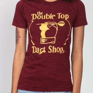 May include: A maroon t-shirt with a gold graphic of a hand throwing a dart at a dartboard. The graphic says "the Double Top Dart Shop."