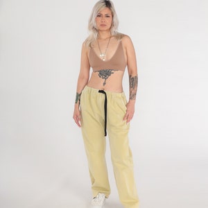 May include: A person wearing a light brown bralette with a gold chain necklace and light yellow pants with a black belt. The person has a tattoo on their chest and arm.