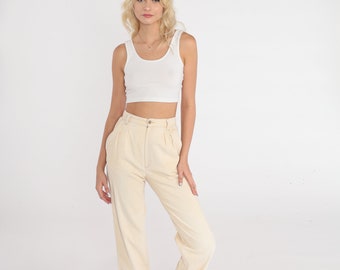 Op de afbeelding: Een vrouw draagt een wit tanktop en een crèmekleurige broek met hoge taille. Ze staat op een witte achtergrond.