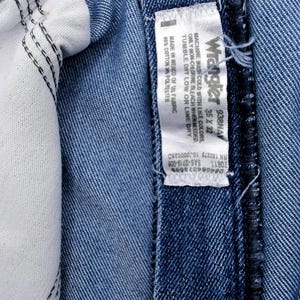 Peut inclure: Gros plan sur un jean en denim bleu, r&eacute;v&eacute;lant les d&eacute;tails int&eacute;rieurs. L'image met en &eacute;vidence la doublure blanche, les coutures et une &eacute;tiquette avec la marque Wrangler et les informations de taille. Le jean est en coton et polyester.