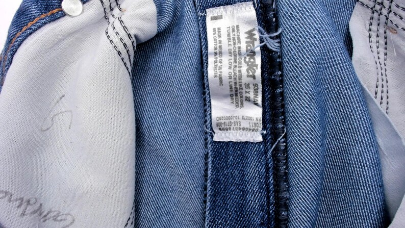 Peut inclure: Gros plan sur un jean en denim bleu, r&eacute;v&eacute;lant les d&eacute;tails int&eacute;rieurs. L'image met en &eacute;vidence la doublure blanche, les coutures et une &eacute;tiquette avec la marque Wrangler et les informations de taille. Le jean est en coton et polyester.
