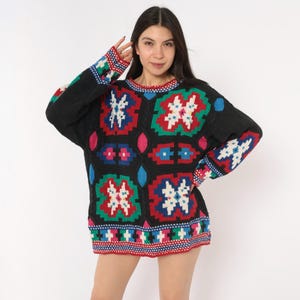 May include: A black long-sleeve jumper featuring a prominent multi-coloured geometric pattern in red, green, blue, and white. The knit garment has a crew neckline, ribbed cuffs, and a repeating block design that extends across the sleeves and body.