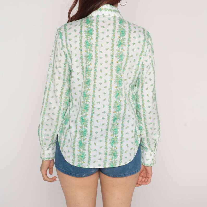Puede incluir: Una camisa blanca de manga larga con botones y un estampado floral verde y blanco. La camisa tiene cuello y un corte holgado.