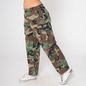 May include: Camouflage cargo pants in shades of green, brown, and black. The pants feature multiple pockets and a classic military-inspired design. The pants are paired with white sneakers.