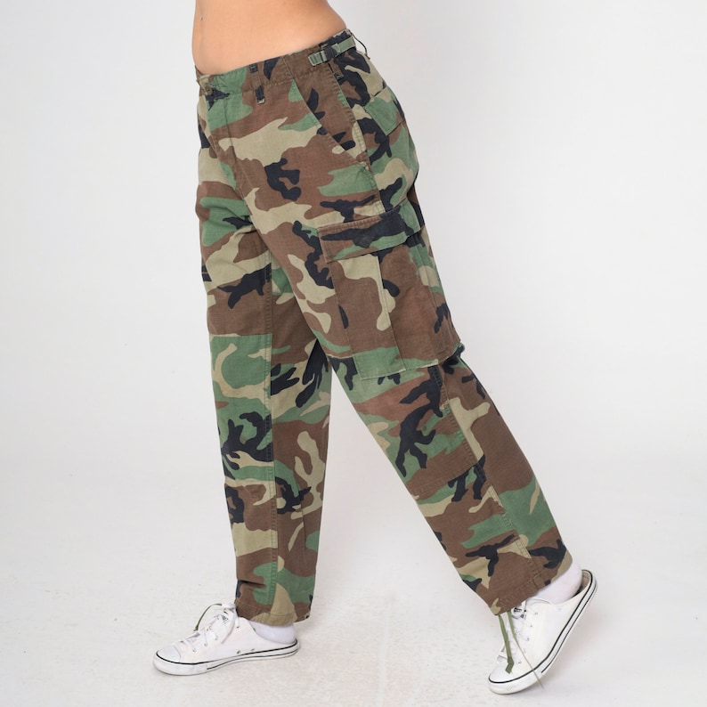May include: Camouflage cargo pants in shades of green, brown, and black. The pants feature multiple pockets and a classic military-inspired design. The pants are paired with white sneakers.