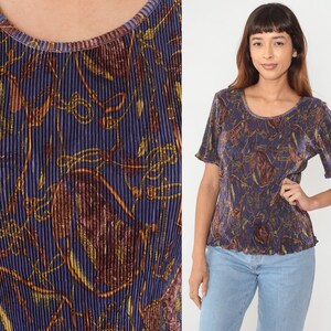 Puede incluir: Una blusa plisada de color morado y dorado con un estampado floral. La blusa tiene mangas cortas y un escote redondo.