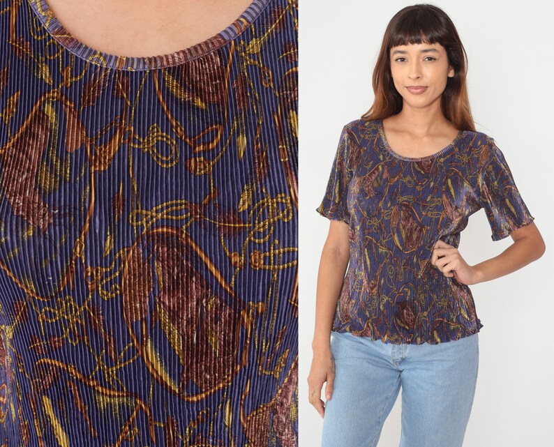Puede incluir: Una blusa plisada de color morado y dorado con un estampado floral. La blusa tiene mangas cortas y un escote redondo.