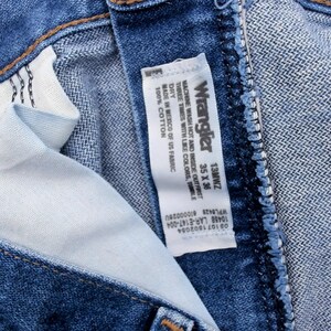 May include: Close-up of blue denim Wrangler jeans. The jeans have a classic five-pocket design and a button and zipper closure. The interior lining is visible, along with the Wrangler label, which includes the size 35 x 36.