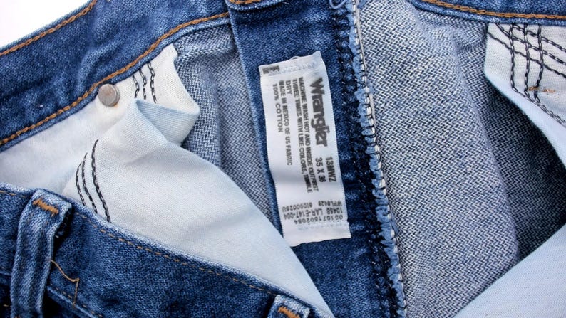 May include: Close-up of blue denim Wrangler jeans. The jeans have a classic five-pocket design and a button and zipper closure. The interior lining is visible, along with the Wrangler label, which includes the size 35 x 36.