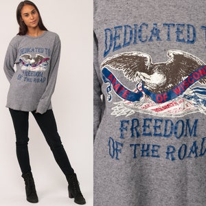 May include: A heather grey long-sleeve shirt featuring a graphic print of an eagle with a banner that reads DEDICATED TO ABATE OF WISCONSIN FREEDOM OF THE ROAD. The shirt has a crew neckline and a textured fabric finish, paired with black pants.
