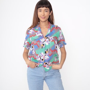 May include: Short-sleeved button-down shirt with a colorful geometric pattern in shades of green, blue, purple, red, and gray. The shirt is paired with light blue denim jeans. The shirt has a collared neckline and is buttoned up.