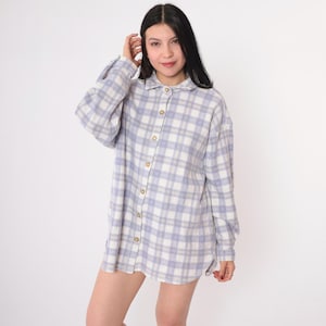 May include: A long-sleeved, oversized plaid shirt in a light blue and white color scheme. The shirt has a button-down front and a relaxed fit.