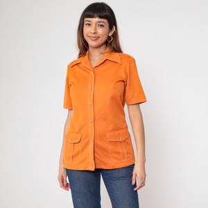 May include: Orange short-sleeved shirt with two front pockets and white stitching. The shirt has a collared neckline and button-up closure. The model is wearing blue jeans.