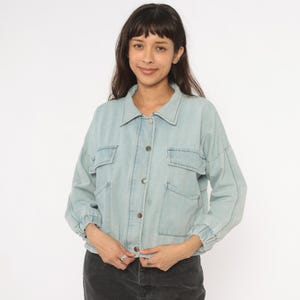 May include: Light blue denim jacket with a button-up front, two chest pockets, and long sleeves. The jacket has a classic collar and a slightly cropped fit. The stitching is visible in a darker shade of blue.