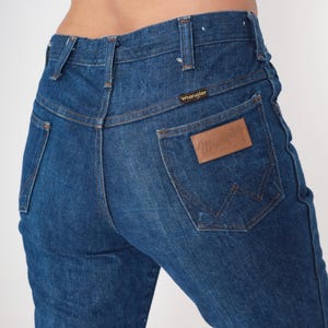 May include: Close-up of blue denim jeans with the Wrangler logo on the back pocket. The jeans have a high waist and classic five-pocket design. The denim is a medium wash, showing some wear.