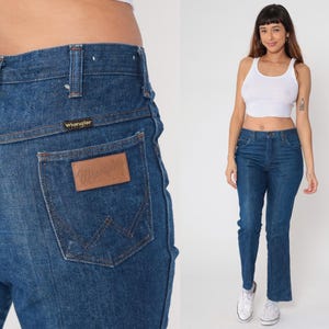 May include: Dark blue denim Wrangler jeans. The jeans have a classic five-pocket design, a straight leg, and a brown leather patch on the back pocket. The Wrangler logo is visible on the patch and above the back pocket.