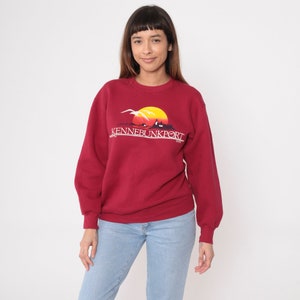 Può includere: Una felpa bordeaux con la scritta "KENNEBUNKPORT" in bianco. La grafica presenta un sole giallo che tramonta su una scena costiera con gabbiani. La felpa è a maniche lunghe e ha un girocollo.