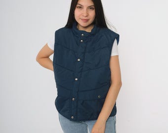 May include: A navy blue quilted vest with a stand-up collar and snap closures. The vest has two front pockets and a quilted design. The model is wearing a white t-shirt and blue jeans.