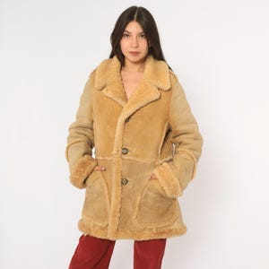May include: A tan suede coat featuring a thick, light-colored shearling lining and trim. The coat has a wide collar, long sleeves with shearling cuffs, two large front patch pockets, and a three-button closure down the center front.