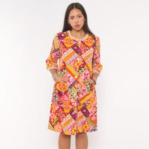May include: A knee-length dress featuring a vibrant patchwork pattern in shades of orange, yellow, pink, purple, and green. The garment has three-quarter length sleeves, a round neckline with white lace trim, a front button closure, and two pockets.