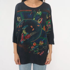 May include: Dark navy blue, oversized t-shirt with a colorful, abstract graphic print. The print features a woman riding a red scooter, surrounded by various food items and geometric shapes. The shirt has three-quarter sleeves.