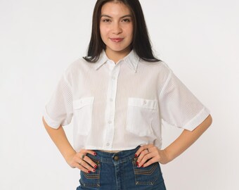May include: White, short-sleeved button-up shirt with two chest pockets. The shirt is paired with high-waisted, dark-wash denim jeans. The shirt has a vertical striped pattern.