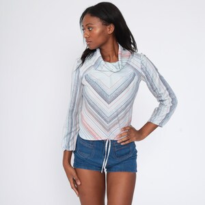 May include: A woman wearing a light blue and pink striped long-sleeve shirt with a cowl neck and a drawstring waist. She is also wearing denim shorts.