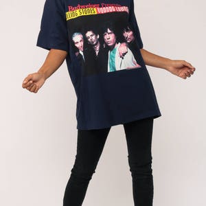 May include: Navy blue t-shirt featuring a graphic of The Rolling Stones band with the text "Budweiser Presents Rolling Stones Voodoo Lounge". The shirt is paired with black jeans and black boots.