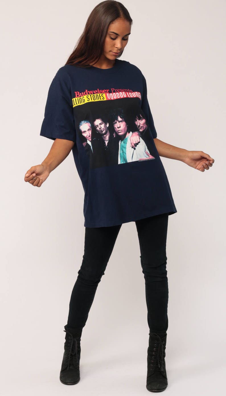 May include: Navy blue t-shirt featuring a graphic of The Rolling Stones band with the text "Budweiser Presents Rolling Stones Voodoo Lounge". The shirt is paired with black jeans and black boots.