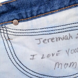 May include: Close-up of the inside of blue denim jeans. The white pocket lining has handwritten text that reads "Jeremiah 29:11 I LOVE You! Mom". The jeans have a metal button and visible stitching.