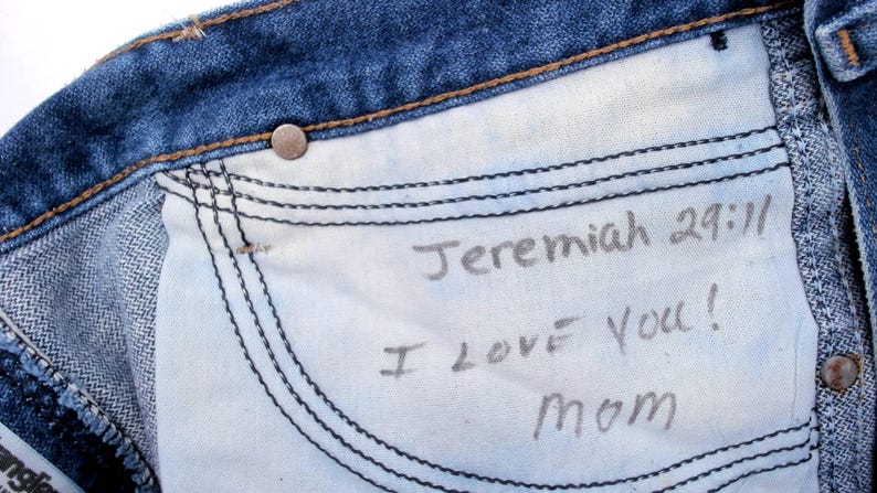 May include: Close-up of the inside of blue denim jeans. The white pocket lining has handwritten text that reads "Jeremiah 29:11 I LOVE You! Mom". The jeans have a metal button and visible stitching.
