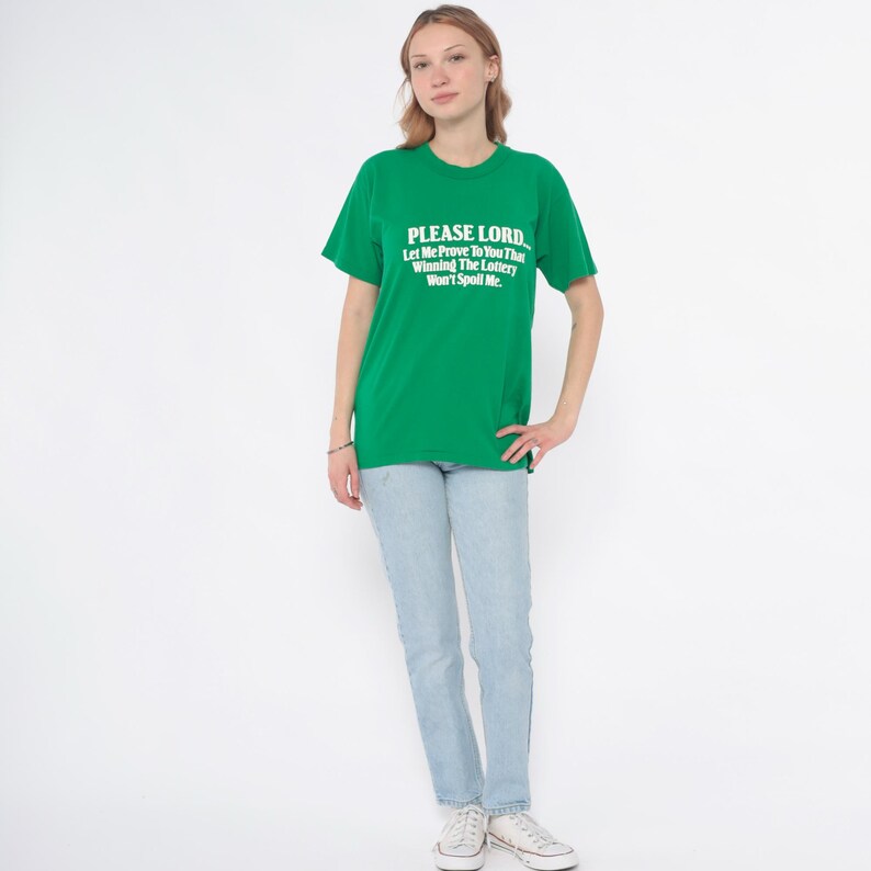 K&ouml;nnte beinhalten: Ein gr&uuml;nes T-Shirt mit dem Text "PLEASE LORD, Let Me Prove To You That Winning The Lottery Won't Spoil Me." Das Shirt wird mit hellblauen Jeans und wei&szlig;en Turnschuhen getragen. Die Person steht vor einem wei&szlig;en Hintergrund.