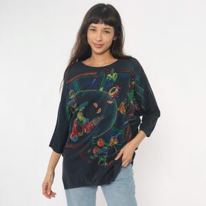 May include: Dark navy blue, oversized top with a colorful graphic print. The print features a scooter, figures, and abstract shapes in red, green, and yellow. The top has three-quarter sleeves and a round neckline.