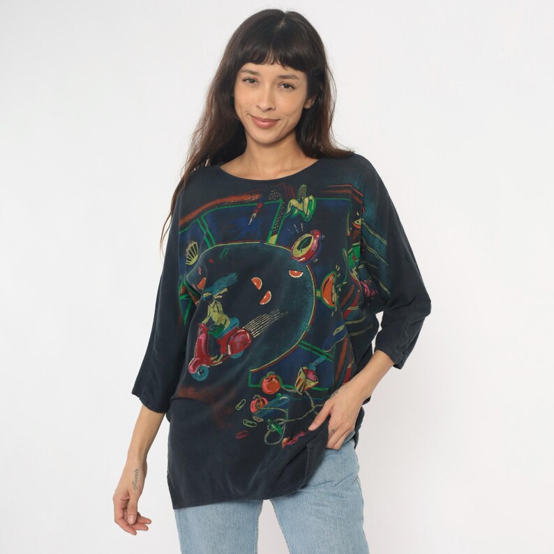 May include: Dark navy blue, oversized top with a colorful graphic print. The print features a scooter, figures, and abstract shapes in red, green, and yellow. The top has three-quarter sleeves and a round neckline.