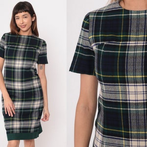 May include: A green, navy, and cream plaid dress with short sleeves. The dress has a straight silhouette and a green hemline. The fabric has a classic plaid pattern with yellow accents. The dress is a vintage style.