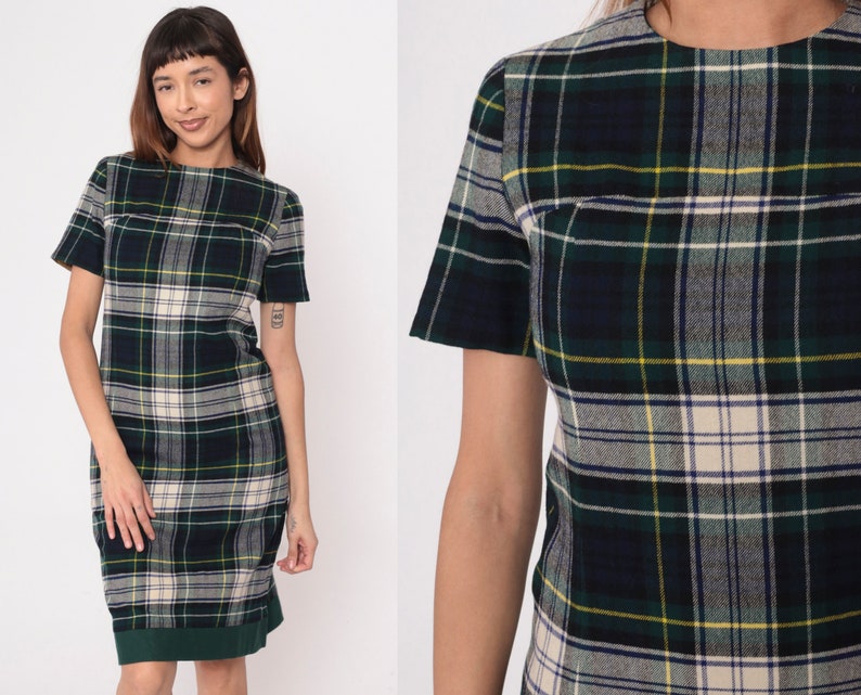May include: A green, navy, and cream plaid dress with short sleeves. The dress has a straight silhouette and a green hemline. The fabric has a classic plaid pattern with yellow accents. The dress is a vintage style.