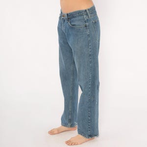 May include: Light blue denim jeans with a classic five-pocket design. The jeans have a straight leg fit and show some wear and tear on the right leg. The jeans are a medium wash and have a button and zipper closure.