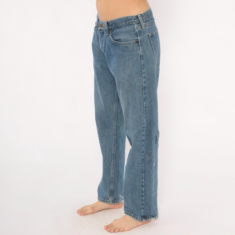 May include: Light blue denim jeans with a classic five-pocket design. The jeans have a straight leg fit and show some wear and tear on the right leg. The jeans are a medium wash and have a button and zipper closure.