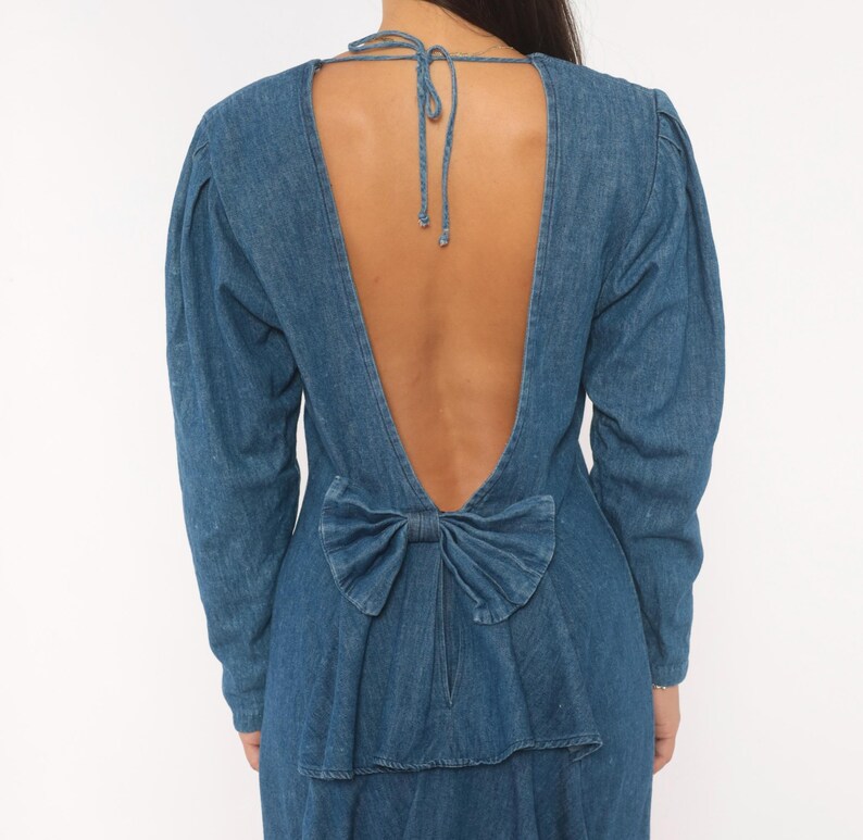 Puede incluir: Un vestido de mezclilla azul de manga larga con un dise&ntilde;o de espalda abierta profunda. La prenda incluye un cierre de lazo en la nuca y un detalle de lazo decorativo de mezclilla en la parte baja de la espalda, sobre una cremallera vertical.