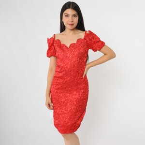 May include: A bright red dress with a textured, crinkled fabric. The dress features a scalloped neckline and short, puffed sleeves. Small embellishments are scattered across the dress. The dress is knee-length and form-fitting.