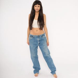 Pu&ograve; includere: Jeans di denim blu chiaro con vestibilit&agrave; rilassata. I jeans hanno un classico design a cinque tasche e una chiusura con bottone e cerniera. La modella indossa un top corto bianco. Lo sfondo &egrave; bianco.