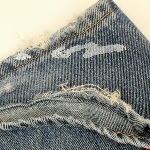 May include: Close-up of a section of blue denim jeans. The fabric is distressed with frayed edges and white paint splatters. The stitching is visible, and the overall look is vintage.