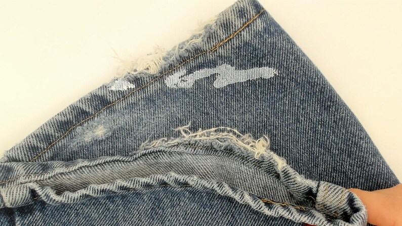 May include: Close-up of a section of blue denim jeans. The fabric is distressed with frayed edges and white paint splatters. The stitching is visible, and the overall look is vintage.