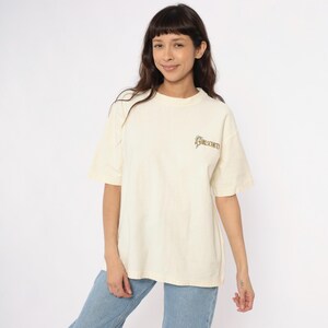 May include: Cream-colored t-shirt with short sleeves and a crew neck. The shirt features the text "GIRLSCOUTS" in a stylized font on the upper right chest. The model is wearing light blue jeans.